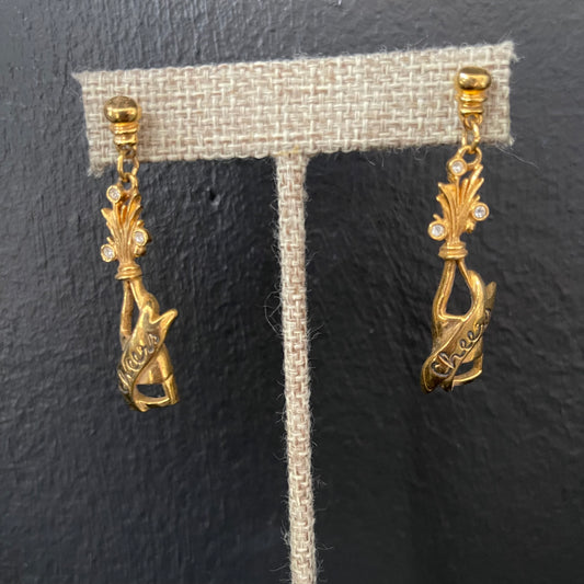 Gold champagne bottle earrings with “Cheers” engraved on them on a gray earring stand against a dark background