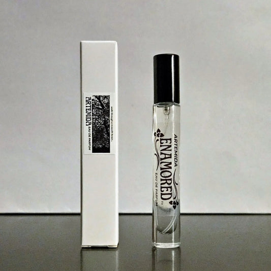 A clear glass perfume roller bottle with a black cap stands next to its tall white box. The bottle and box are labeled 'ARTEMIDA ENAMORED EAU DE PARFUM' with floral graphics, set against a neutral gray background.
