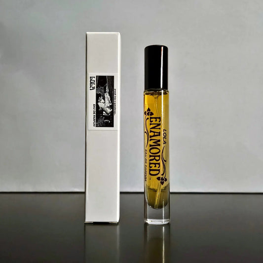 A clear cylindrical perfume bottle filled with golden-yellow liquid, with a black cap, standing next to its tall white rectangular box. The bottle is printed with the words 'LOLA ENAMORED EAU DE PARFUM' and decorative black graphics.