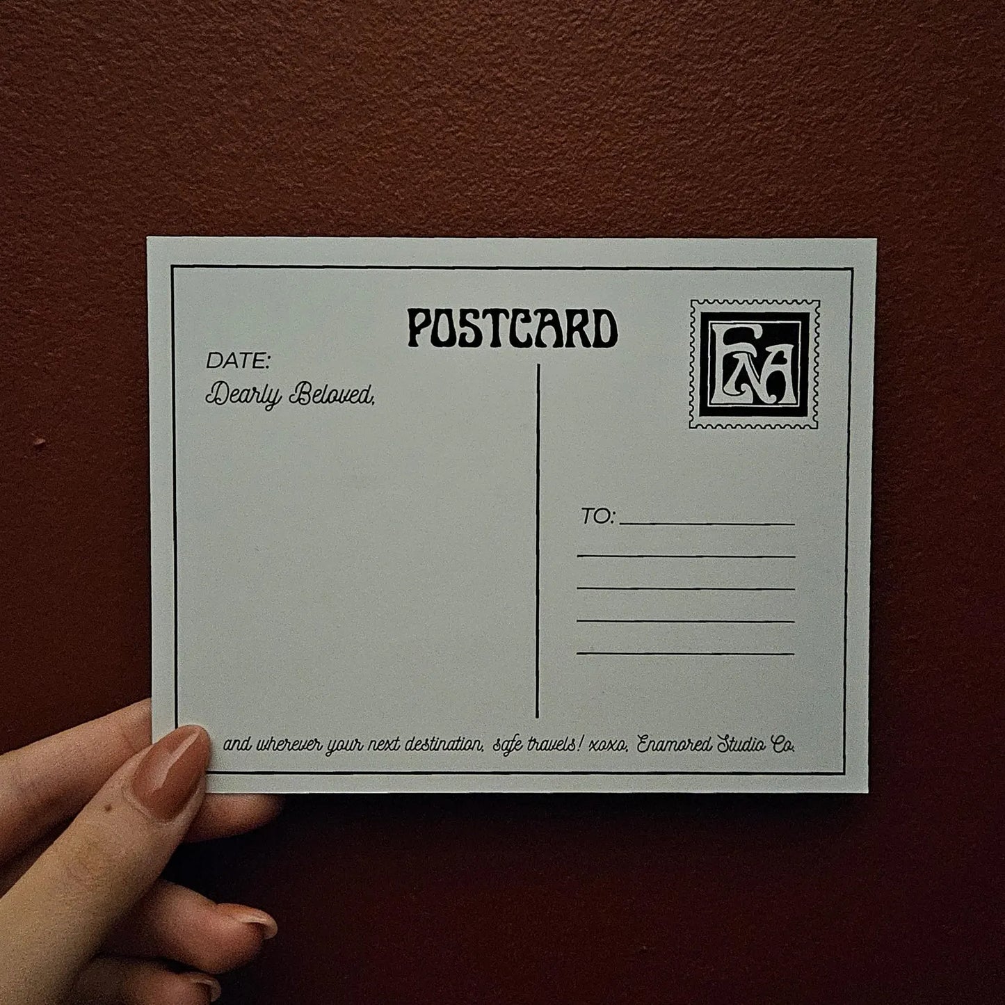 A hand with manicured nails holds a white postcard against a textured dark red background. The postcard features black printed text fields for date, destination, and a decorative stamp graphic.