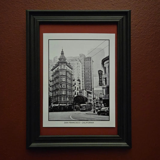 A framed black and white postcard with a photograph of a San Francisco street scene, mounted on a textured reddish-brown wall. The print features a white mat, a red inner mat, and a thick black frame.