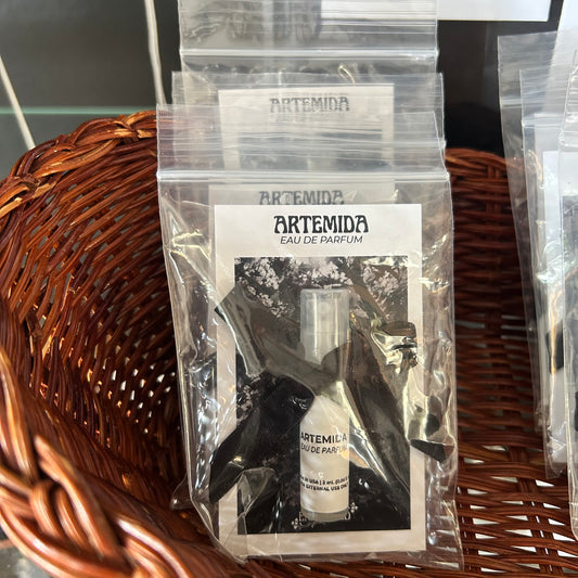 A clear plastic resealable pouch containing a small perfume bottle and a printed card labeled ARTEMIDA EAU DE PARFUM, placed on a brown woven wicker surface.