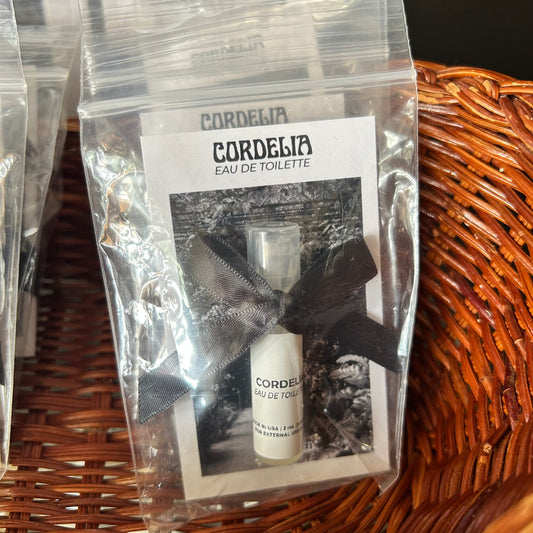 A small clear plastic pouch containing a miniature white perfume sample bottle tied with a dark ribbon, placed on a brown woven wicker surface. The insert card reads CORDELIA EAU DE TOILETTE.