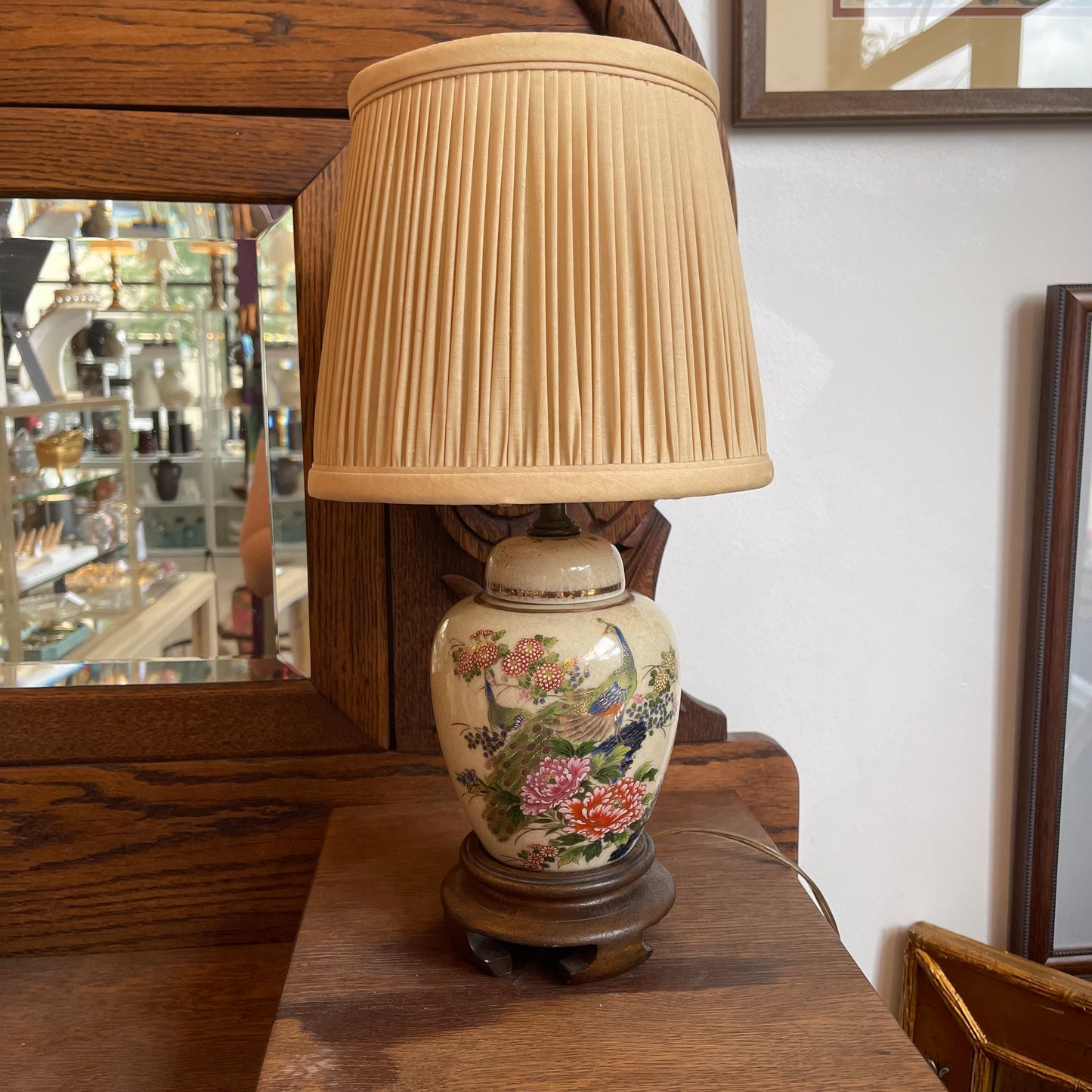 A vintage-style table lamp with a pleated beige fabric shade and a ceramic base decorated with colorful floral and bird motifs, set on a wooden surface in an indoor room.