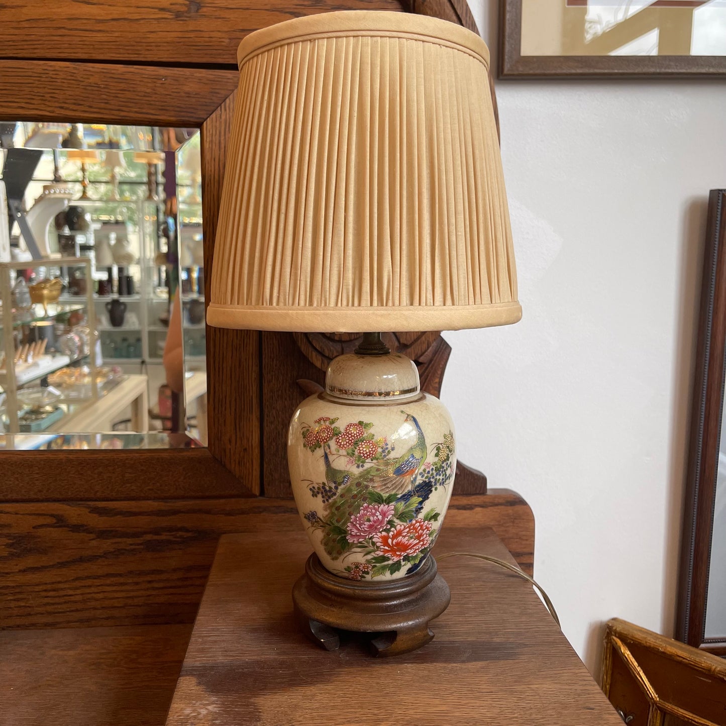 A vintage-style ceramic table lamp with a pleated beige fabric shade and a hand-painted floral base, displayed on a wooden surface in a shop setting.