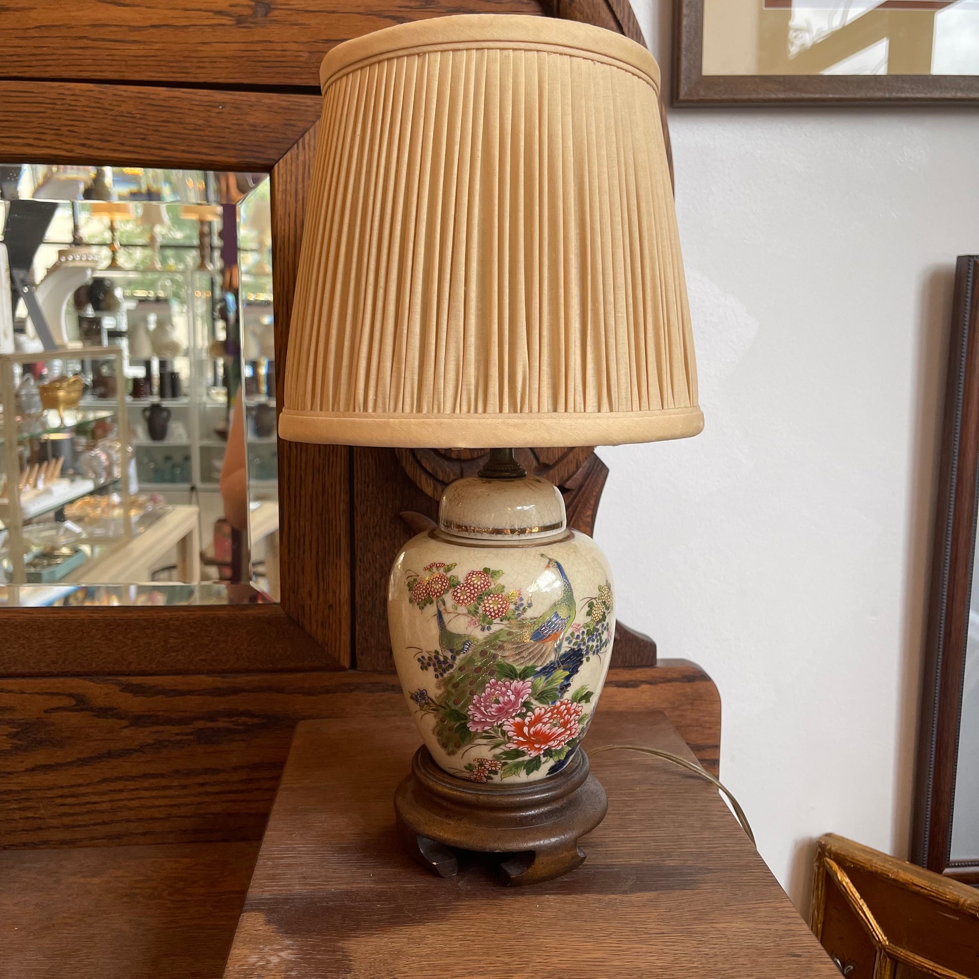 A vintage-style ceramic table lamp with a pleated beige fabric shade and a hand-painted floral base, displayed on a wooden surface in a shop setting.