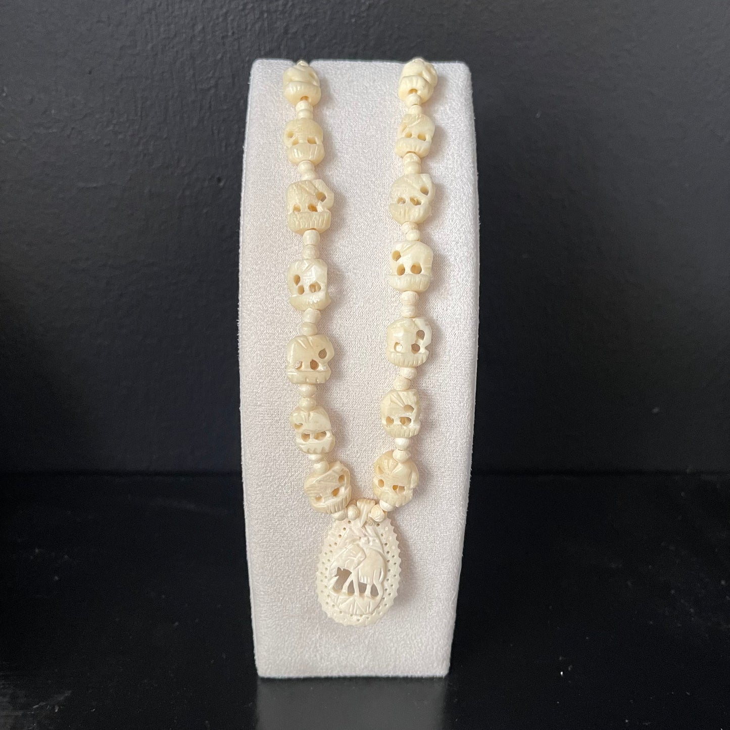 Necklace with ivory elephant beads with carved elephant pendant on a white stand against a dark background