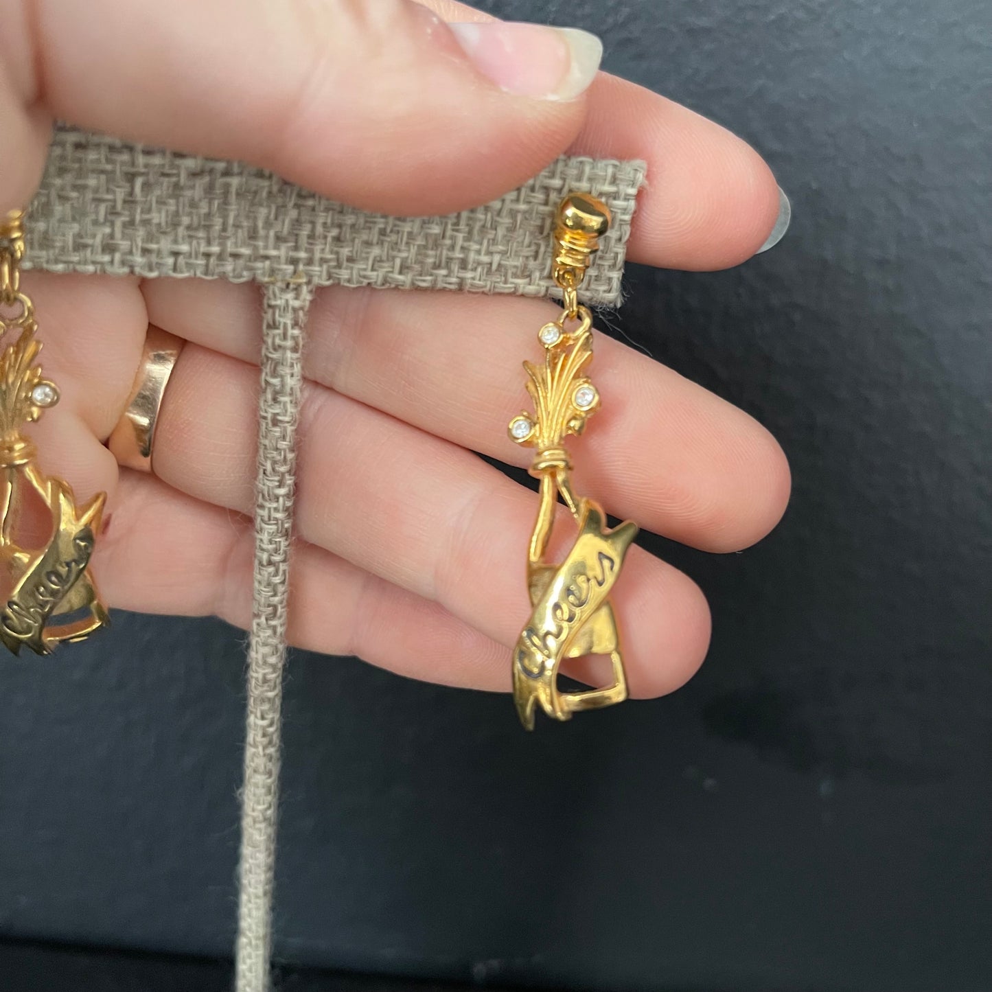 Gold champagne bottle earrings with 'Cheers' engraved on them, held by a hand against a dark background.
