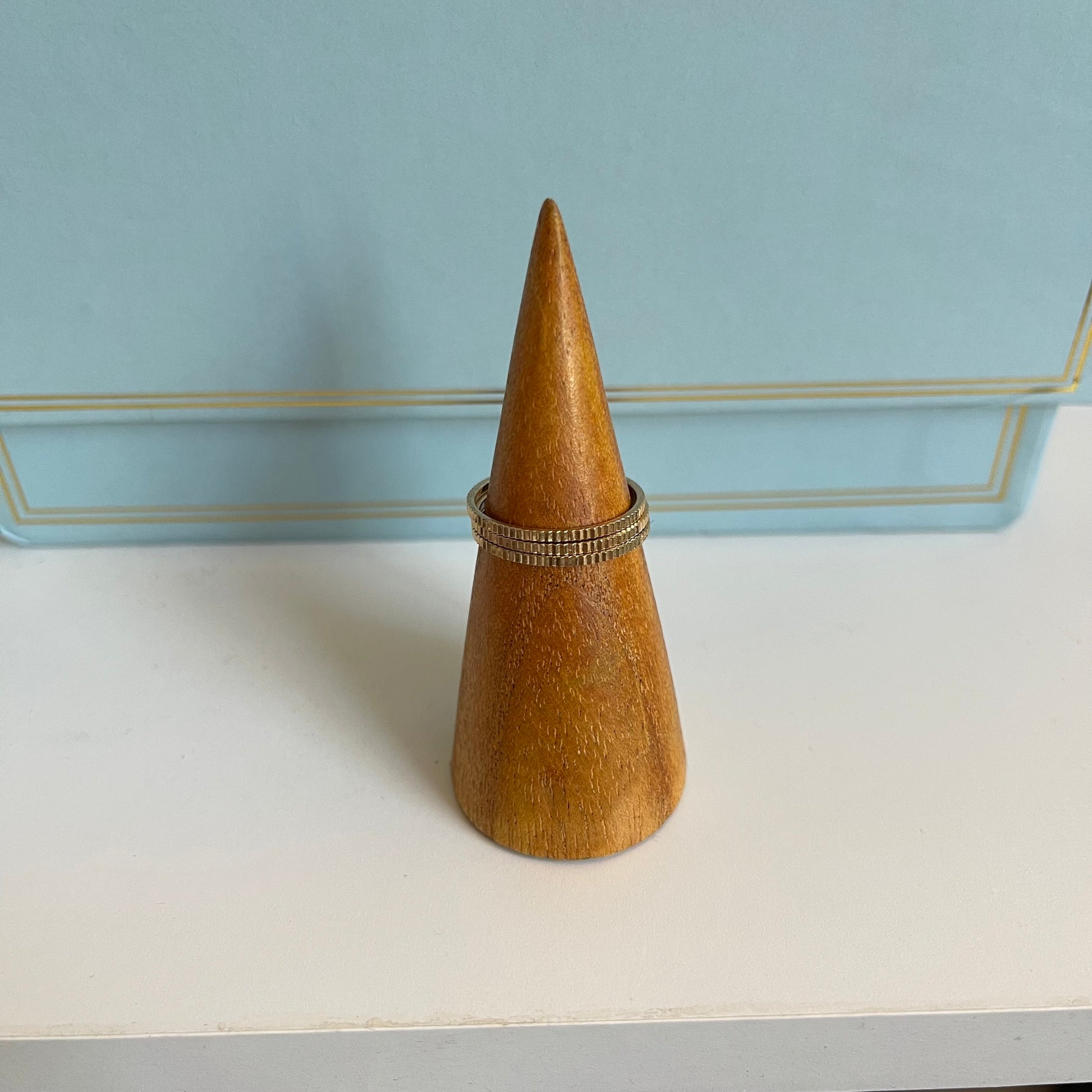 Wooden cone-shaped ring holder with 3 thin gold ridged rings on a light blue background