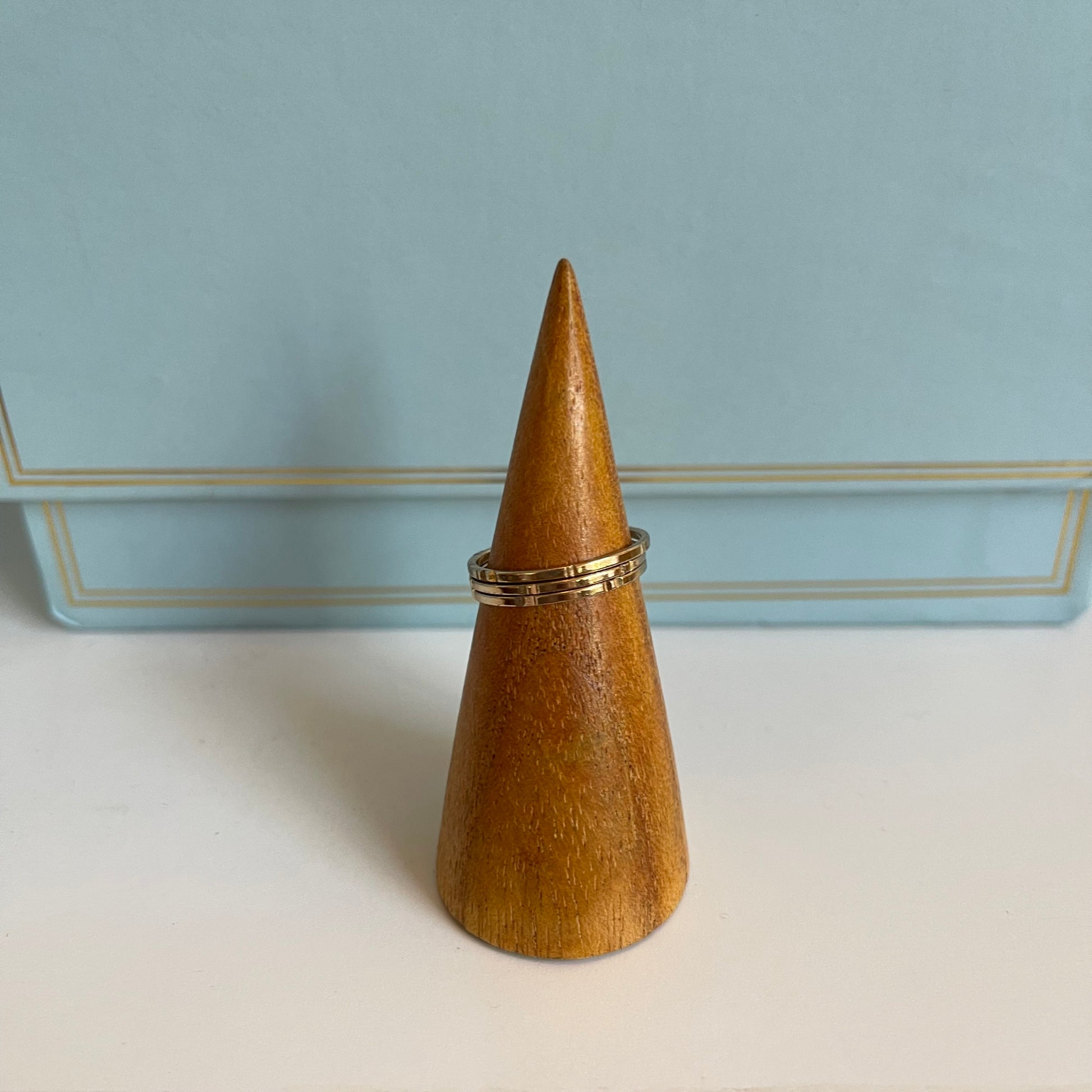 Wooden cone-shaped ring holder with 3 thin gold rings on a light blue background