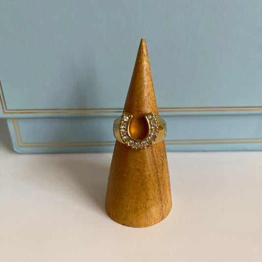 Gold horseshoe ring with crystals and thick band on wooden cone-shaped ring display against light blue background 