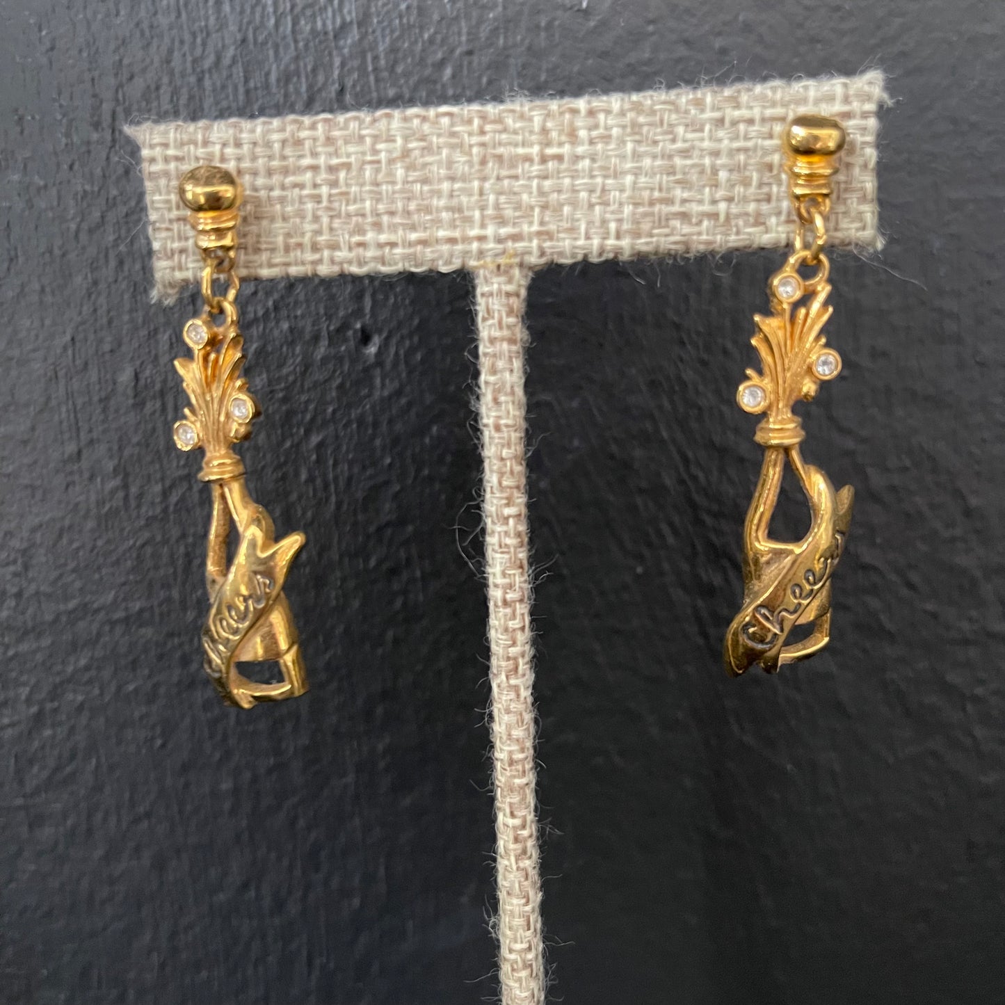 Gold champagne bottle earrings with “Cheers” engraved on them on a gray earring stand against a dark background