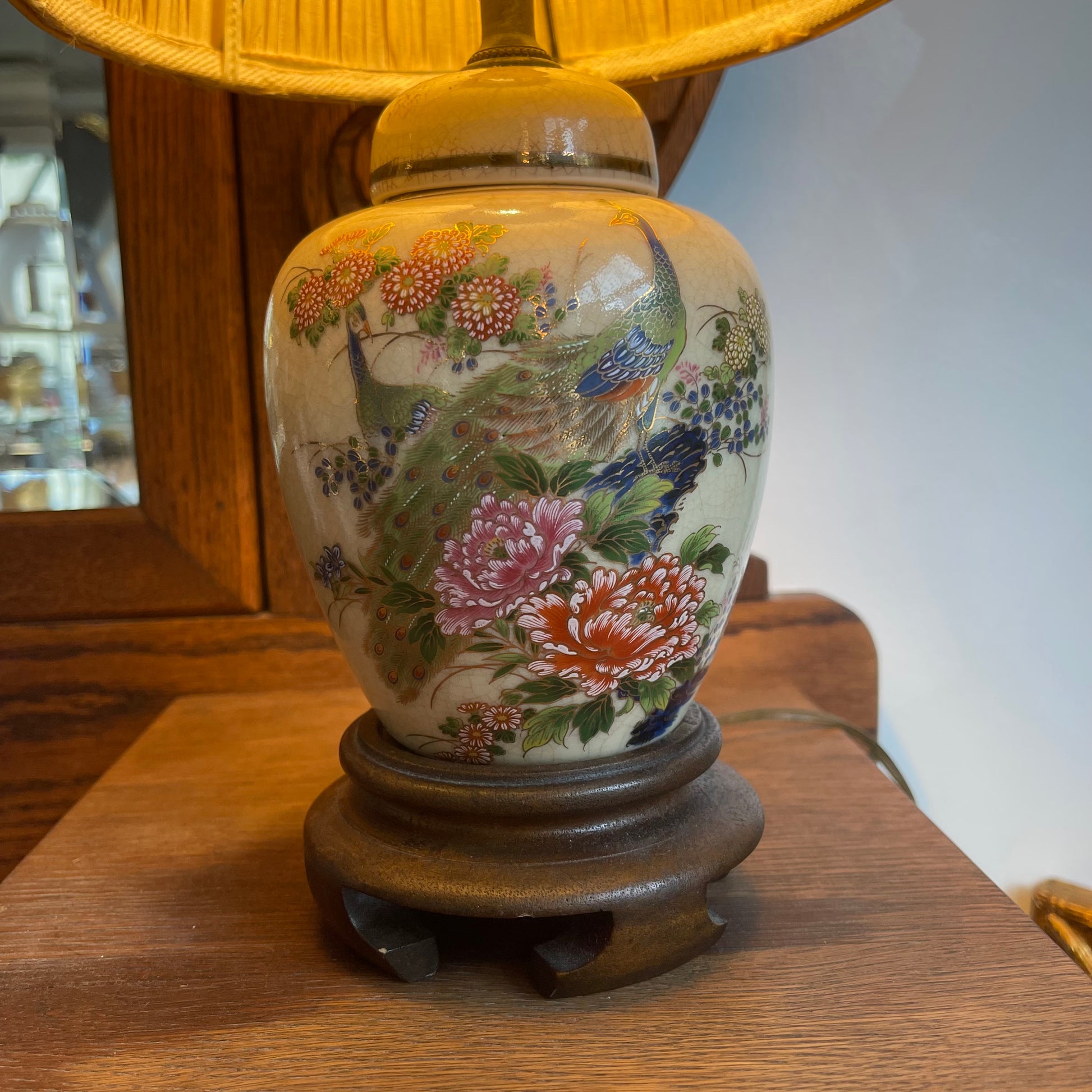 A ceramic table lamp with a cream-colored body decorated with colorful hand-painted peacocks and flowers, resting on a dark wooden base next to a yellow fabric lampshade.