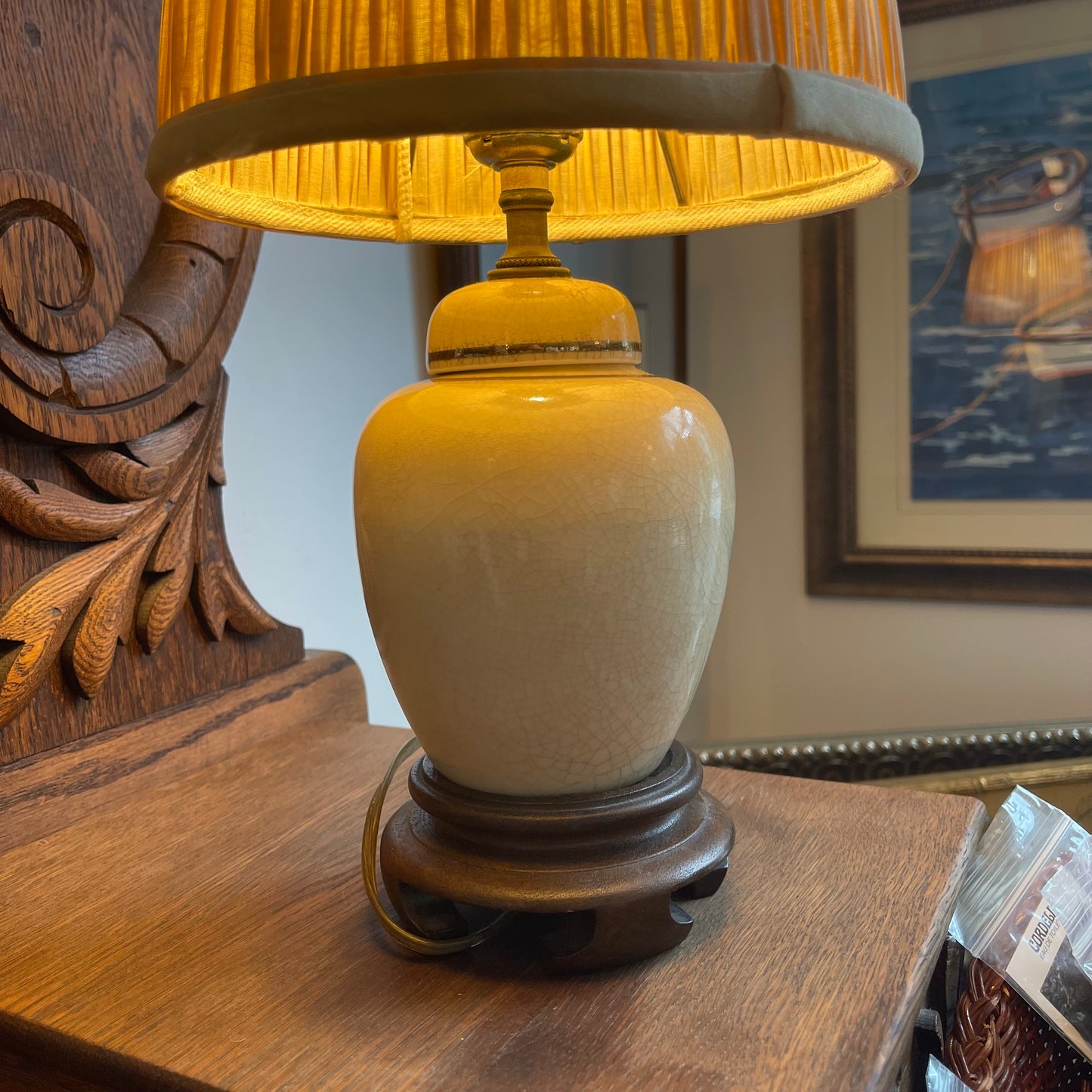 A ceramic table lamp with a warm yellow-gold shade, a cream-colored glazed jar-shaped body, and a dark brown wooden base, shown lit on a wooden table in a home setting.