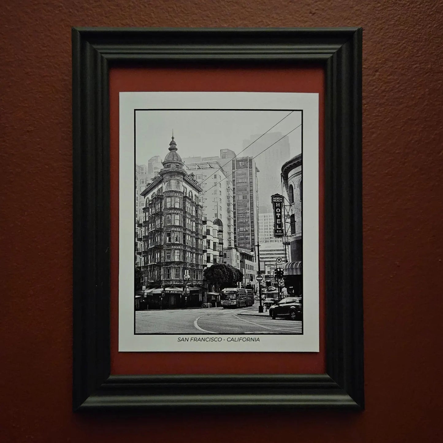 A framed black and white postcard with a photograph of a San Francisco street scene, mounted on a textured reddish-brown wall. The print features a white mat, a red inner mat, and a thick black frame.