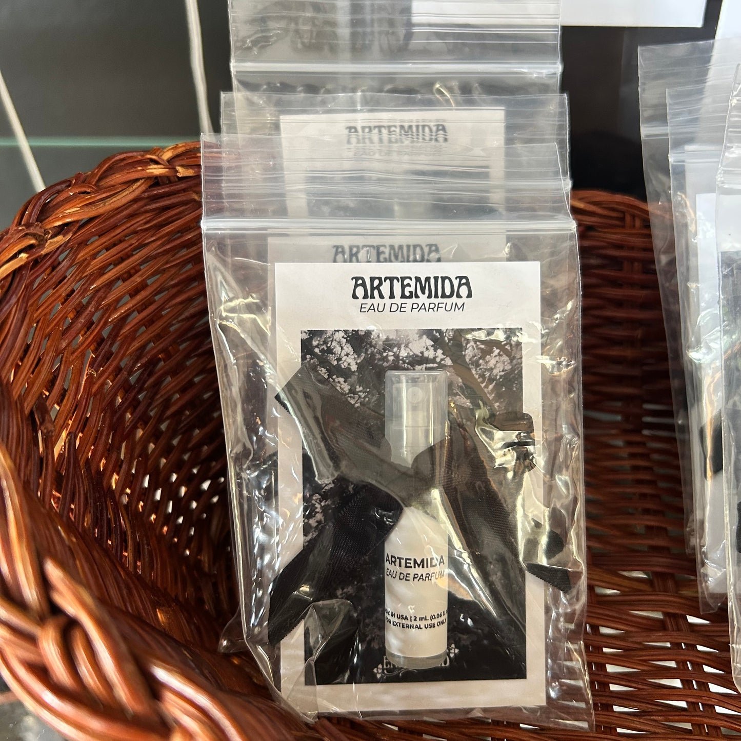 A clear plastic resealable pouch containing a small perfume bottle and a printed card labeled ARTEMIDA EAU DE PARFUM, placed on a brown woven wicker surface.