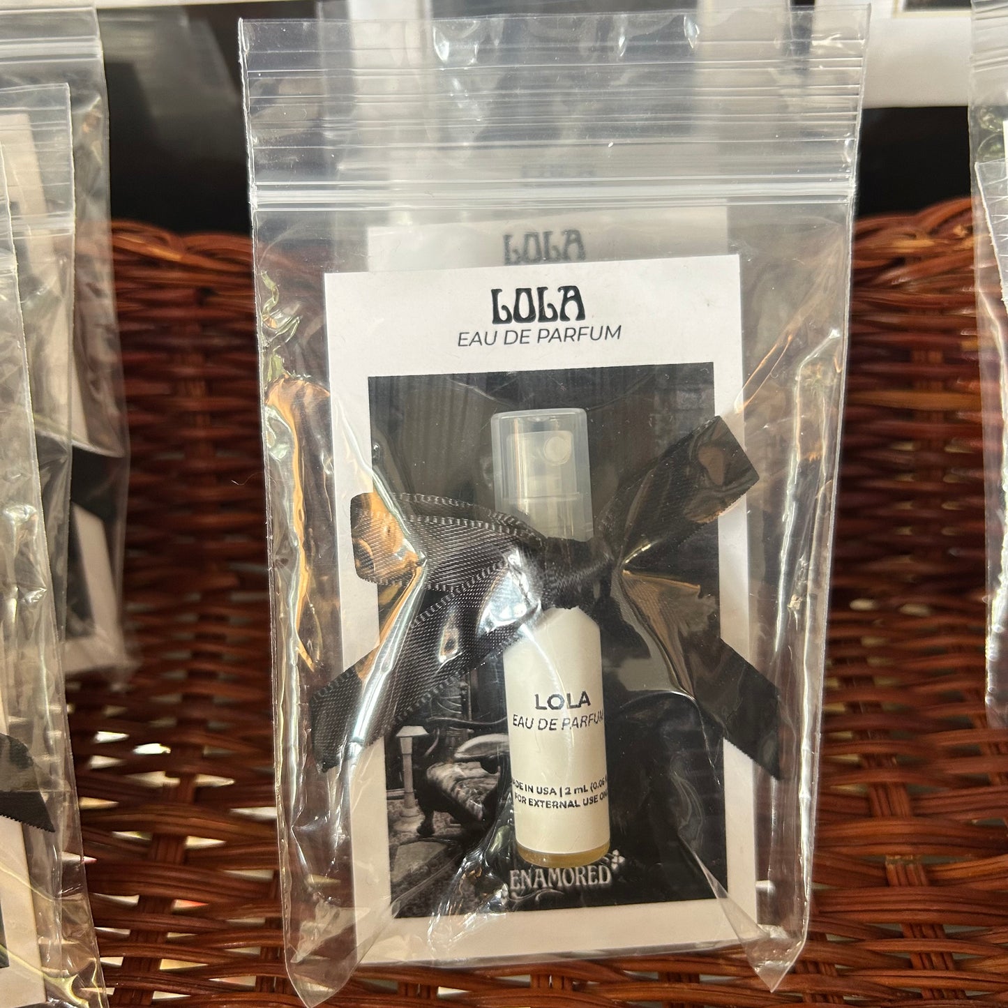 A small perfume sample vial wrapped in a black ribbon and presented inside a clear plastic bag with a printed card reading 'LOLA EAU DE PARFUM'. The vial is placed on a brown woven wicker surface.